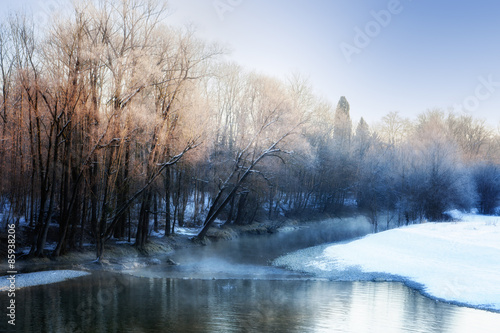 Small river in winter, with sunbeams filtering through bare birch trees. Winter landscape. Winter wonderland. Winter background.