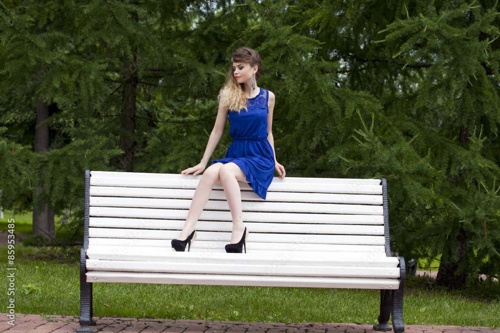 Beautiful blonde girl in blue dress sitting on a bench in summer