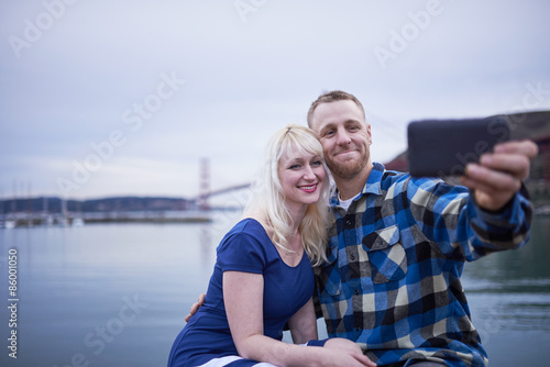 couple sitting by water front beside golden gate bridge taking selfies together © Joshua Resnick