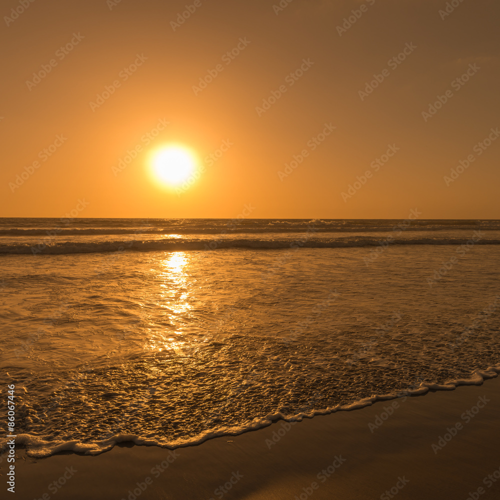 Sun sets over horizon of ocean. Surf on sandy beach. Sky, water and shore in the evening painted in orange, gold color. Sun sets over horizon of ocean. Surf on sandy beach. Sky, water and shore in the evening painted in orange, gold color.
