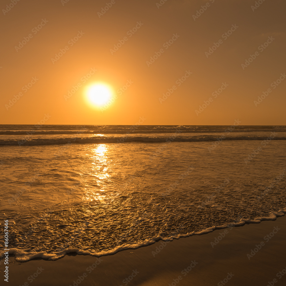 Sun sets over horizon of ocean. Surf on sandy beach. Sky, water and shore in the evening painted in orange, gold color.