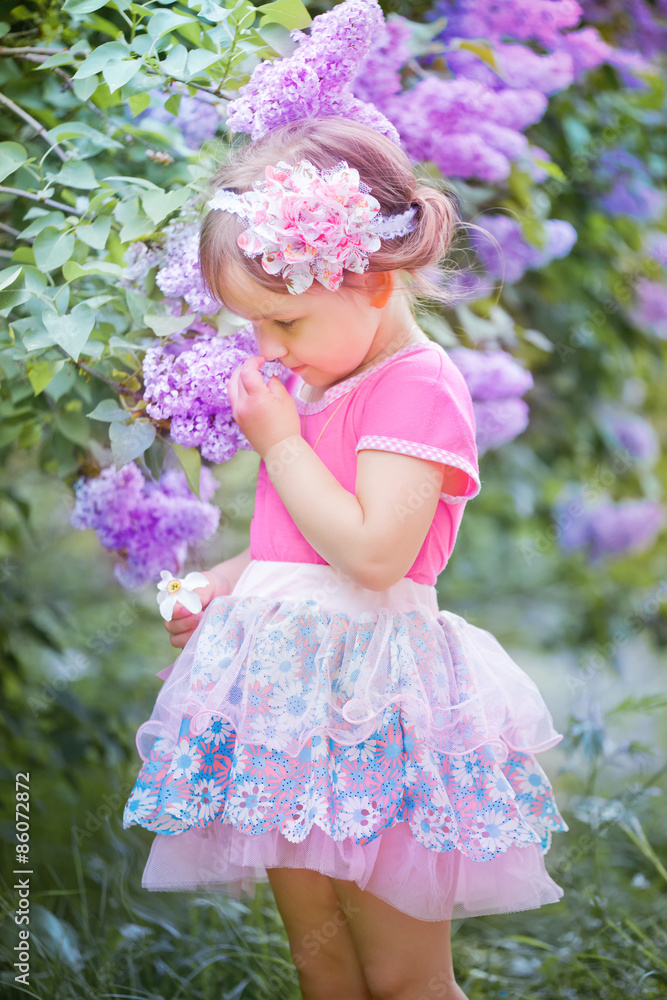 Fototapeta premium Little beautiful girl standing in the lilac park on a sunny morning and smiling.