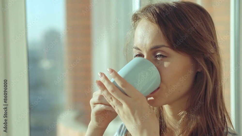 Young woman drinking coffee by the window during sunrise in her cozy home. Relaxed morning at home concept. Slow motion filmed at 240 fps. Young woman drinking coffee by the window during sunrise in her cozy home. Relaxed morning at home concept. Slow motion filmed at 240 fps.