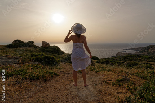 Junge Frau mit weisem Kleid und Hut steht im Gegenlicht der Sonne in einer Landschaft mit Bergen und Meer in der Ferne