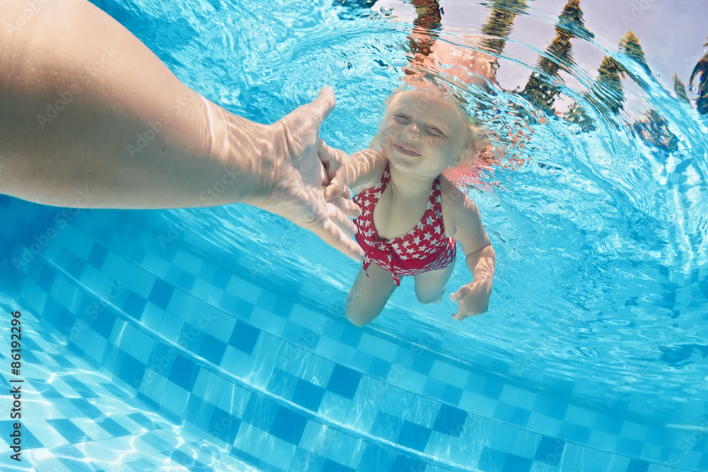 Joyful baby girl diving underwater with fun and holding parents hand for assistance in swimming pool. Healthy active family lifestyle, children water sport activity with mother on summer vacation