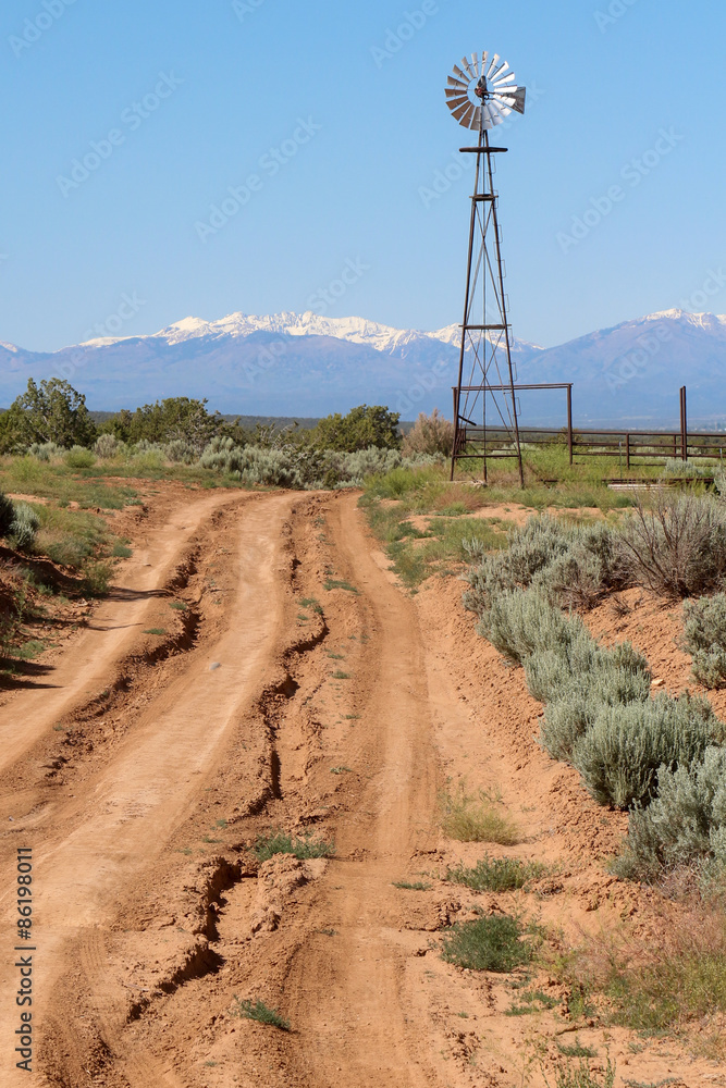 Rutted dirt road leading to a metal windmill and snow covered mountains in New Mexico