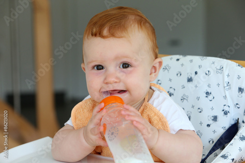 Baby girl drinking and playing with his milk bottle  sitting in a high chair.