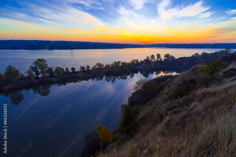 Gorgeous dawn on blue lake.
Luminous sunrise blooming over wild waterfront landscape with fall grass flower hill stone rocks and narrow island with colored trees in the middle of water surface
