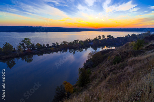 Gorgeous dawn on blue lake. Luminous sunrise blooming over wild waterfront landscape with fall grass flower hill stone rocks and narrow island with colored trees in the middle of water surface
