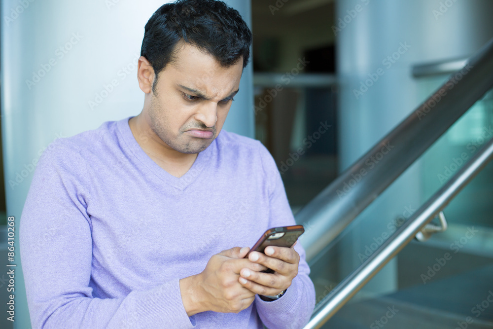 Closeup portrait, stressed young man in purple sweater, shocked surprised, horrified disturbed, by what he sees on his cell phone, isolated indoors office background. Closeup portrait, stressed young man in purple sweater, shocked surprised, horrified disturbed, by what he sees on his cell phone, isolated indoors office background.