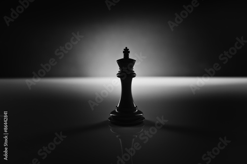 Solitary black chess king carved in genuine ebony wood in focus standing on a glossy table in the dark