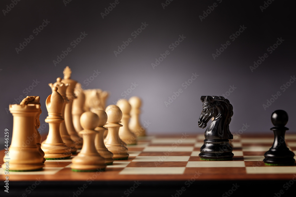 Black chess knight and pawn facing the entire army of white chess pieces on a wooden chessboard in focus Black chess knight and pawn facing the entire army of white chess pieces on a wooden chessboard in focus