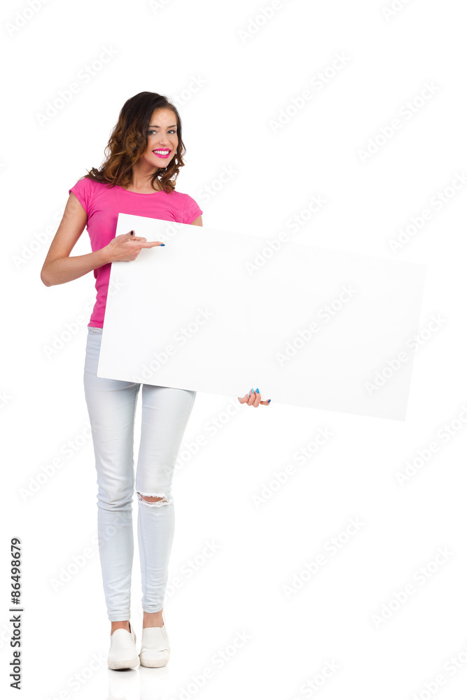 Smiling young woman in torn jeans and pink shirt standing, holding white placard and pointing. Full length studio shot isolated on white Smiling young woman in torn jeans and pink shirt standing, holding white placard and pointing. Full length studio shot isolated on white