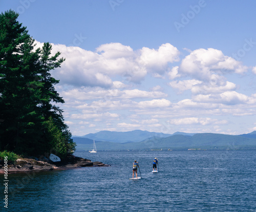 two people enjoying paddle boarding on a lake with a sailboat and mountains in the distance