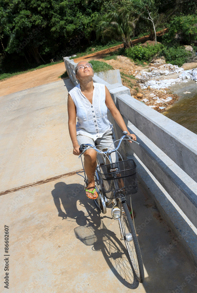 Young woman riding bicycle across river bridge next to tropical park