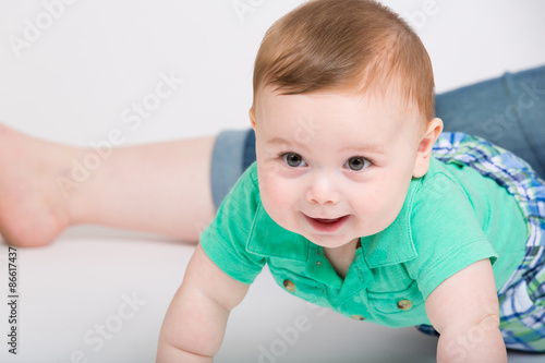 8 month year old baby crawls towards camera, while mom leg is sitting in background. dressed in a cute green polo shirt and blue plaid shorts.