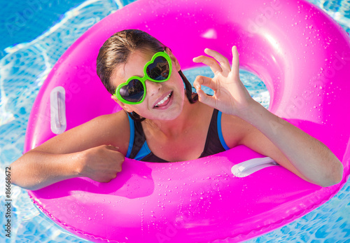 happy teenager playing on a pink buoy in a pool with sunglasses