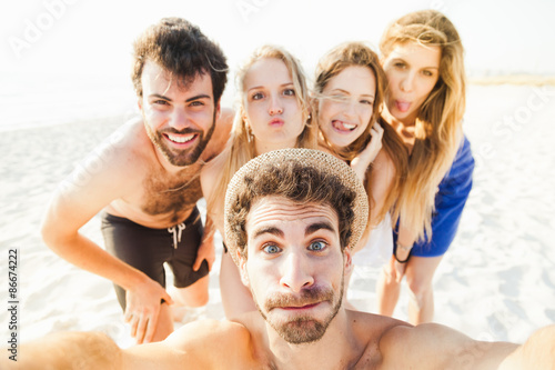 Selfie on the beach! Five friends in swimwear (two men and three women) take a photo at the sea in a summer day at sunset on the beach with no other people and making funny faces 