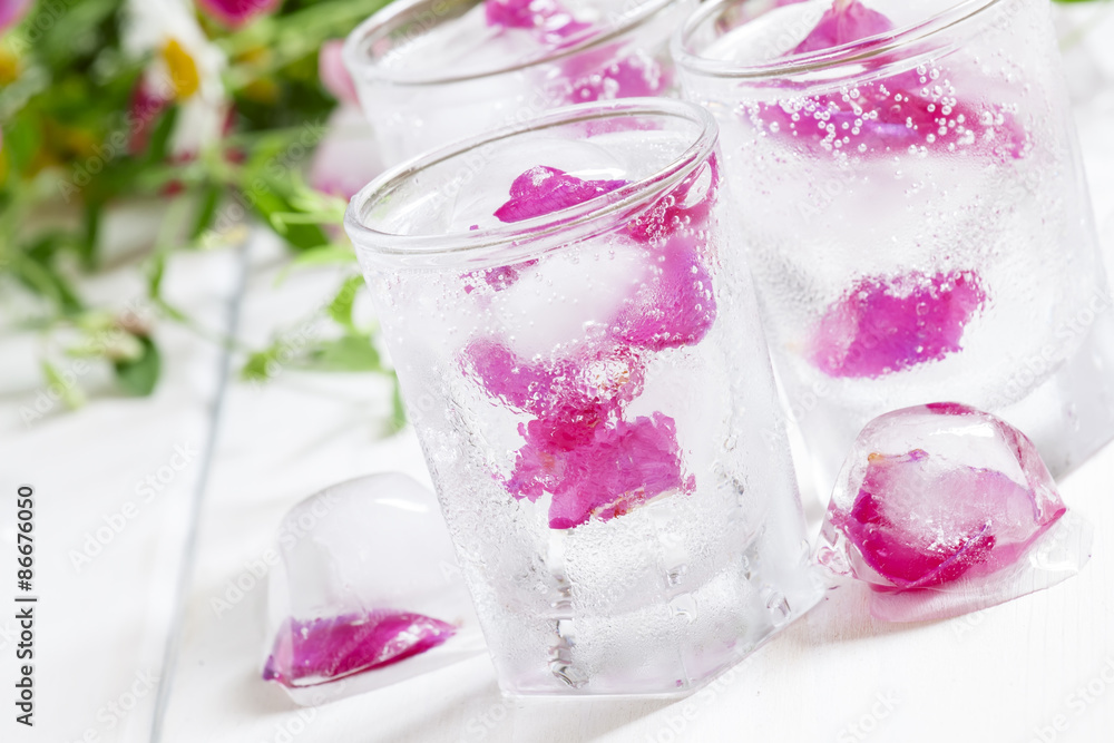 Fresh sparkling beverage with ice with rose petals on white wood