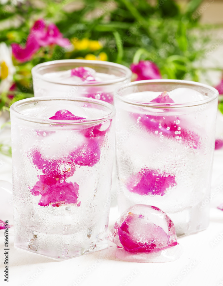 Fresh sparkling beverage with ice with rose petals on white wood