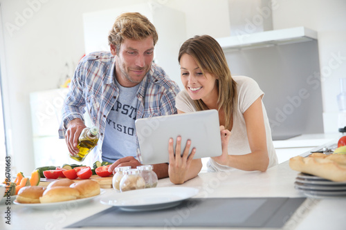 Tableau sur toile Cheerful couple in kitchen cooking dinner, using tablet
