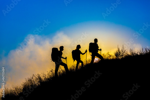 Family journey wild landscape Silhouettes of three people walking with backpacks hiking gear up toward top of wild grass mountain mother father daughter bright luminous sunrise sky background