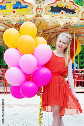 Positive emotions concept, cheerful young woman holding colorful latex balloons in the amusement park in front of the carousel