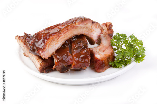 Plate of Barbecued Spareribs – Barbecued, grilled pork spareribs on a white plate. Parsley on the right side. On a white background.