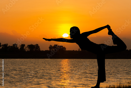 Silhouette young woman practicing yoga at orange sunset on the river background