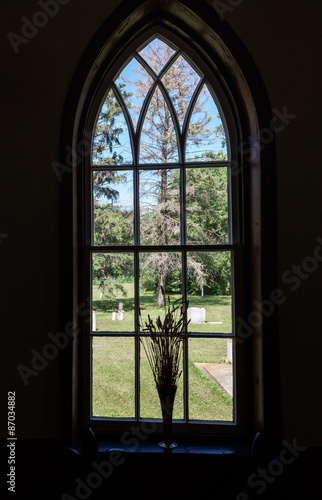 vertical image of an old arched church window with vintage design looking out onto a grave yard under a bright blue sky and green trees in summer time