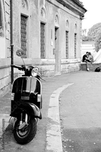 Black and white photo of an iconic scooter with a young couple in the background. #87046889