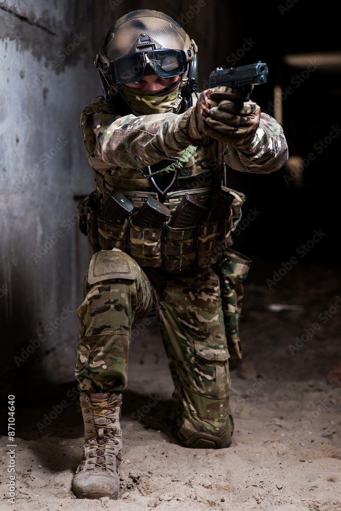 Fototapeta premium Armed man in military camouflage aiming his gun on knees/Man in military ammunition stay on his knees with gun in hand
