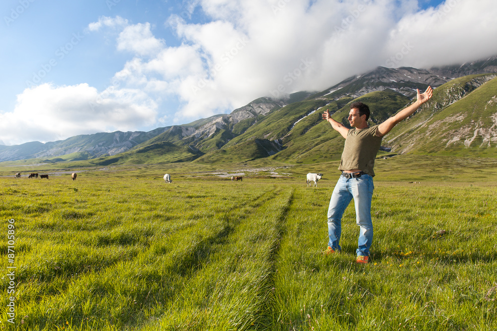 Uomo  alza le braccia al cielo, in un campo d'erba, con montagne e vacche sullo sfondo