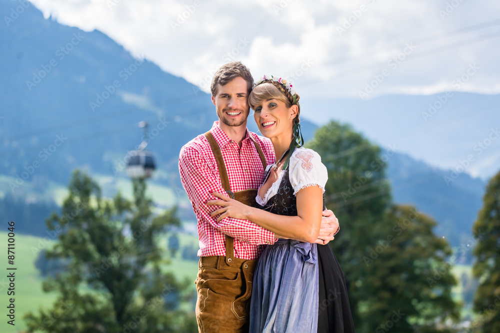 paar-in-tracht-steht-auf-alm-in-den-alpen-vor-einer-seilbahn-in-die