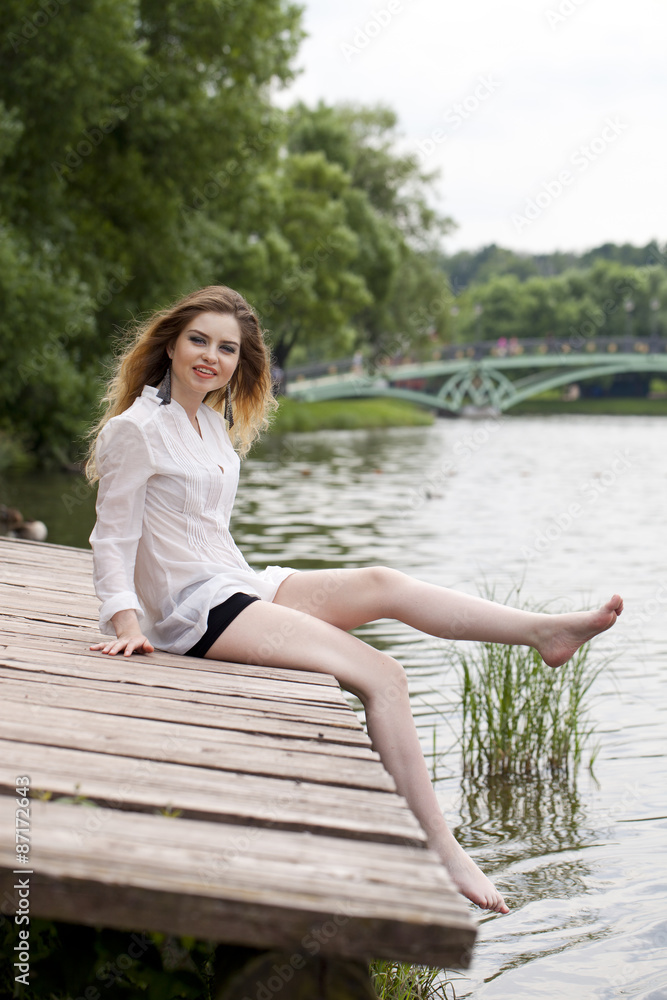Young beautiful girl in a white tunic sits on a wooden pier on t