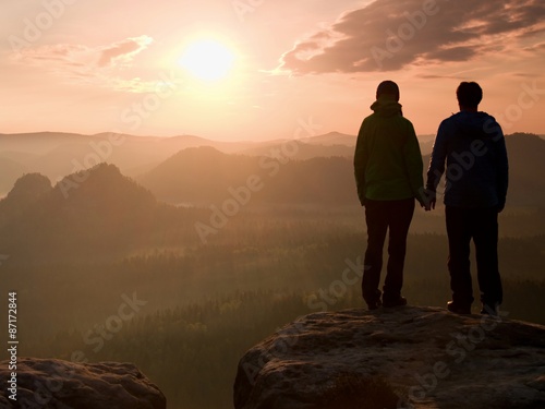 Young pair of hikers hand in hand on the peak of rock empires park and watch over the misty and foggy morning valley to Sun. Beautiful moment the miracle of nature