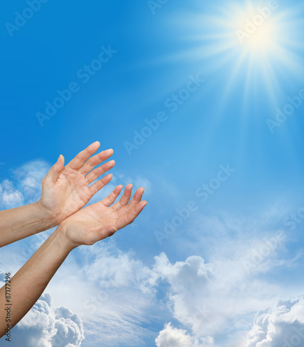 Divine Source - Female hands reaching up towards a bright sun burst on a blue sky background with fluffy clouds and plenty of copy space