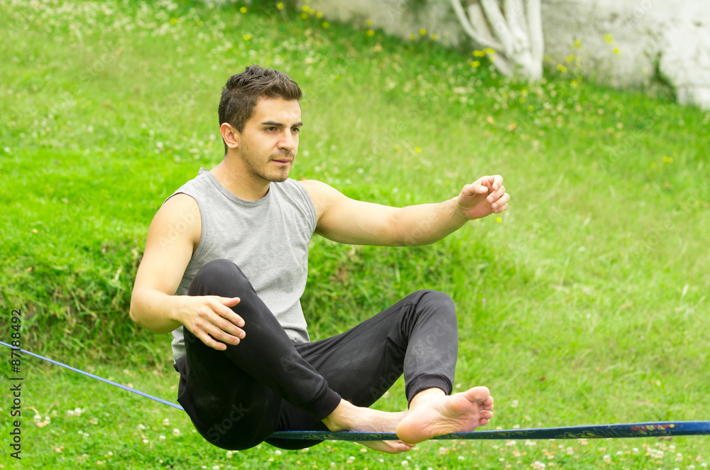 Man sitting on slackline left arm to the side concentrating keeping balance with grassy background Man sitting on slackline left arm to the side concentrating keeping balance with grassy background