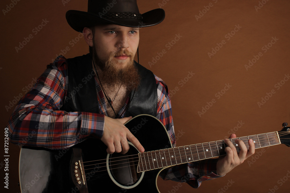 The young handsome bearded man in a leather cowboy's hat and a leather biker jacket with a guitar in hands