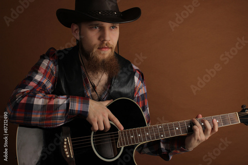 The young handsome bearded man in a leather cowboy's hat and a leather biker jacket with a guitar in hands