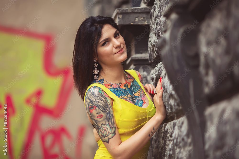 Naklejka premium A beautiful woman in a yellow dress, tattoos, leaned on architec
