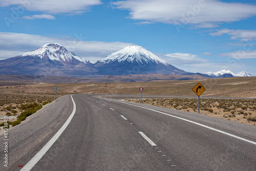 Road running towards the snow and ice covered peaks of the volcanoes Parinacota (6342m) and Pomerape (6240m) rising above the Altiplano of Northern Chile in Lauca National Park. © JeremyRichards