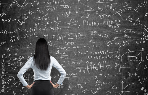 Rear view of a thoughtful woman who tries to solve math problems. Math calculations on black chalk board. photo