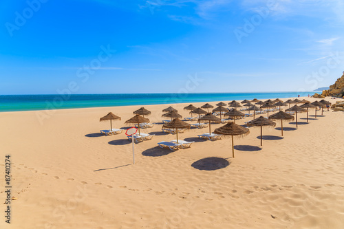 Sunbeds with umbrellas on sandy beach in Salema seaside town, Alrgarve region, Portugal