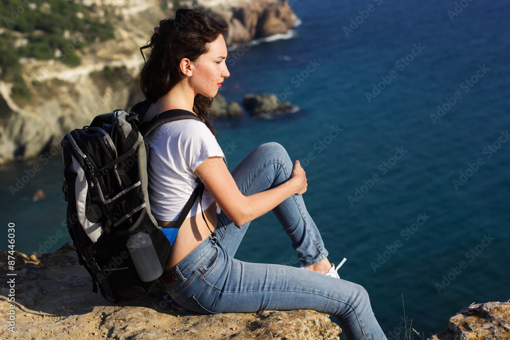 Pretty girl traveler is sitting on rock edge with backpack