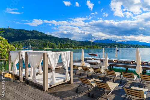 Sunchairs and beds on wooden deck and view of beautiful alpine lake Worthersee in summer time, Austria
