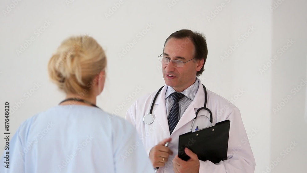Serious doctor with glasses holding a clipboard and giving arrangements to a blonde female colleague, back turned. Indoors Serious doctor with glasses holding a clipboard and giving arrangements to a blonde female colleague, back turned. Indoors