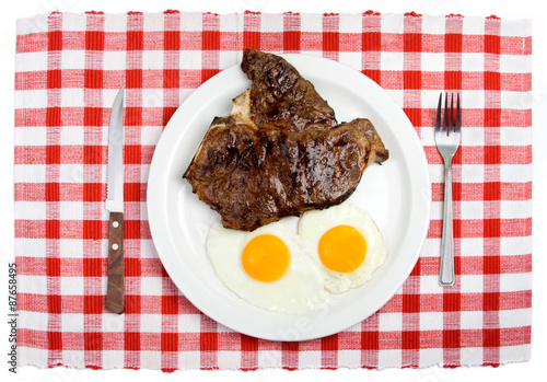 T-Bone Steak and Fried Eggs Breakfast – Two sunny side up eggs and a T-Bone steak on a white plate. Table setting, with fork and steak knife. Red checked background.