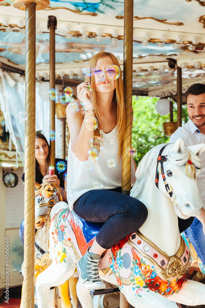 On a summer day a group of friends having fun in the shade on a carousel with horses and blowing soap bubbles. In the foreground a young woman in the air while blowing many colorful soap bubbles