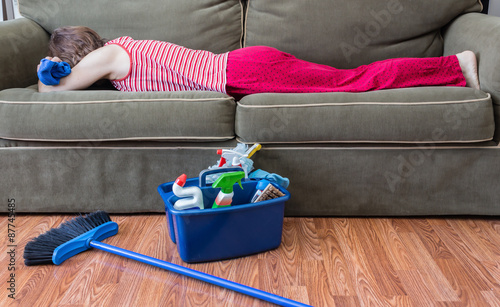 horizontal image of a house keeper sleeping on the couch with a rag in her hand and a broom and housecleaning supplies sitting on the floor next to her